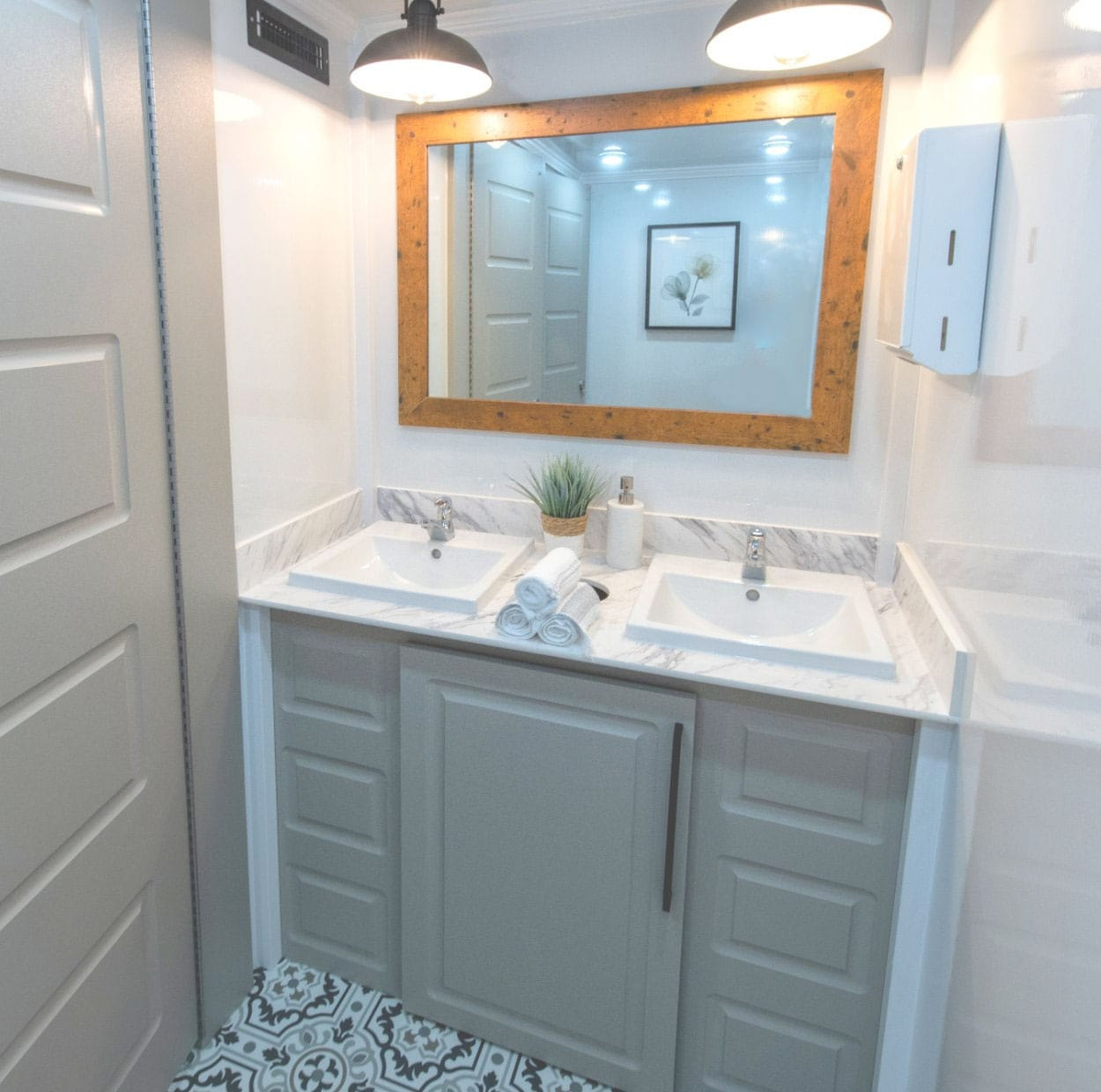 Luxury restroom trailer interior with double sinks, a marble-look counter, gray cabinets and a wood-framed mirror
