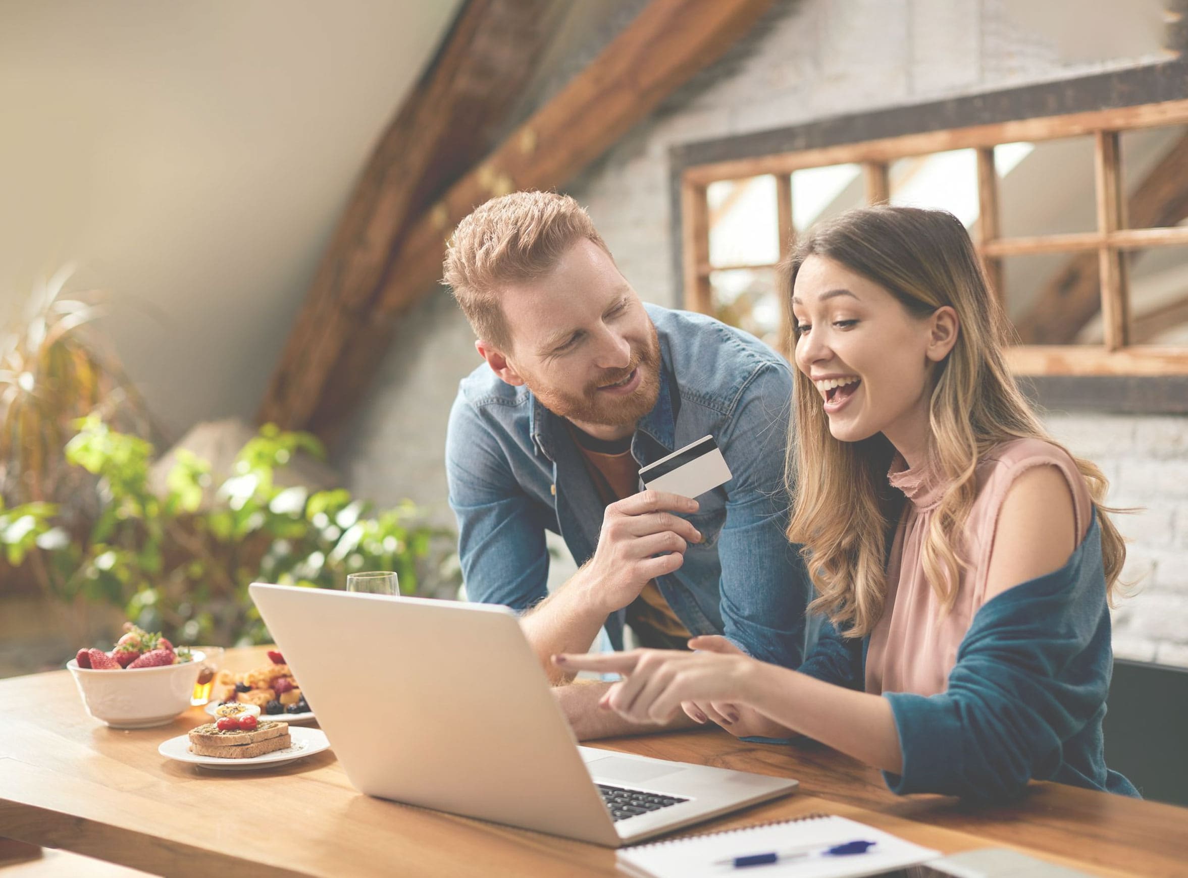 Smiling couple at a kitchen table, one holding a credit card while the other points at a laptop screen