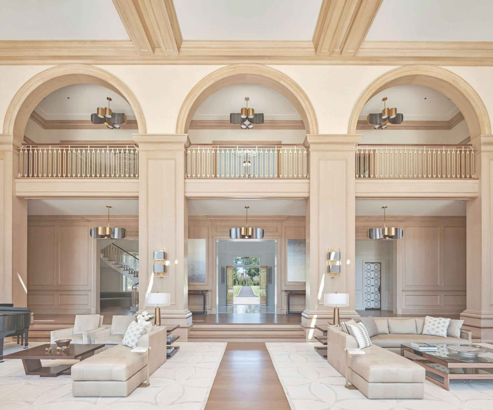 Grand two-story living hall with arched balconies, pale oak paneling, brass chandeliers and two matching cream seating areas